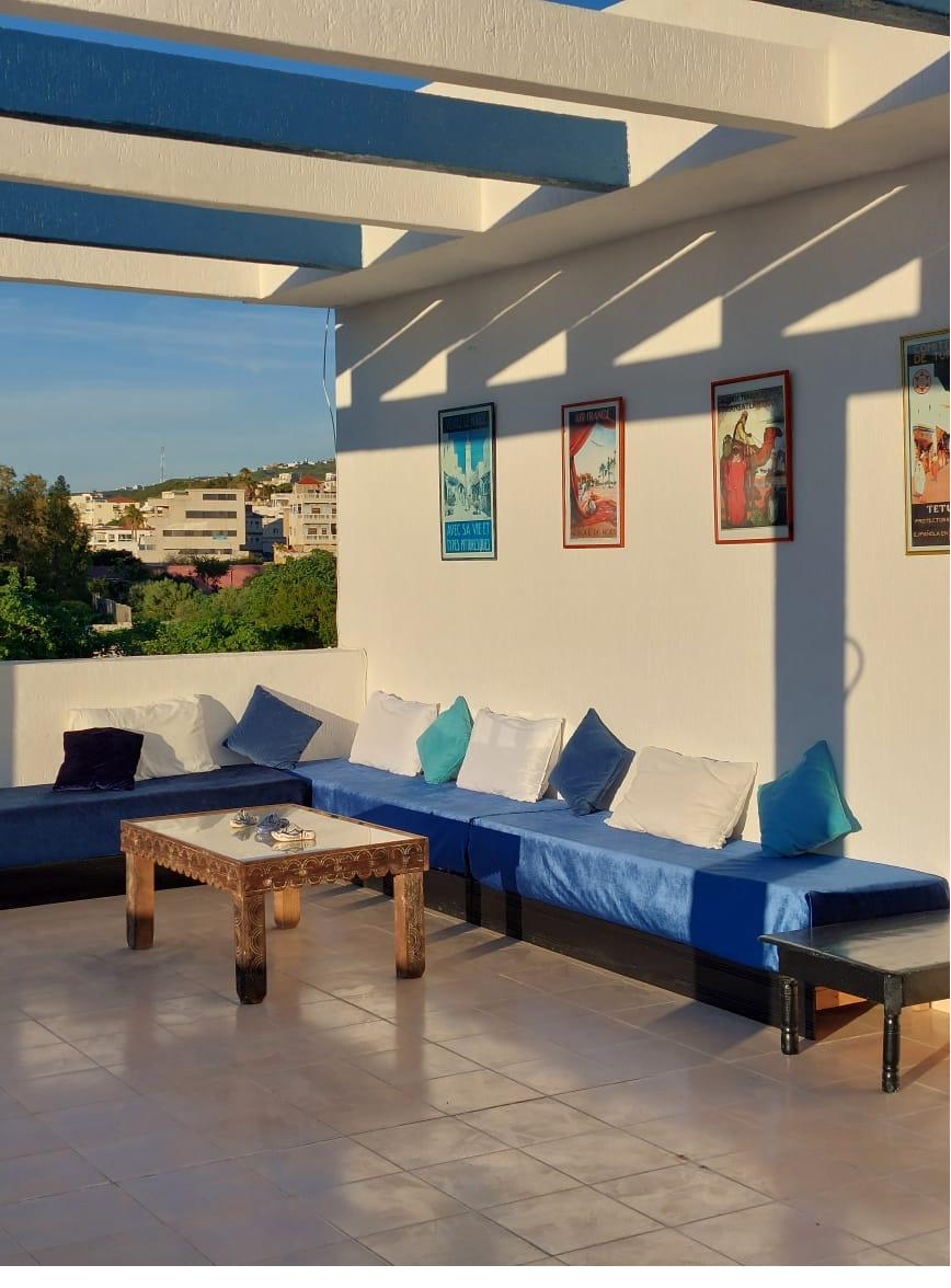Salon du rooftop avec banquettes bleues, coussins, table basse marocaine et affiches vintage du Maroc